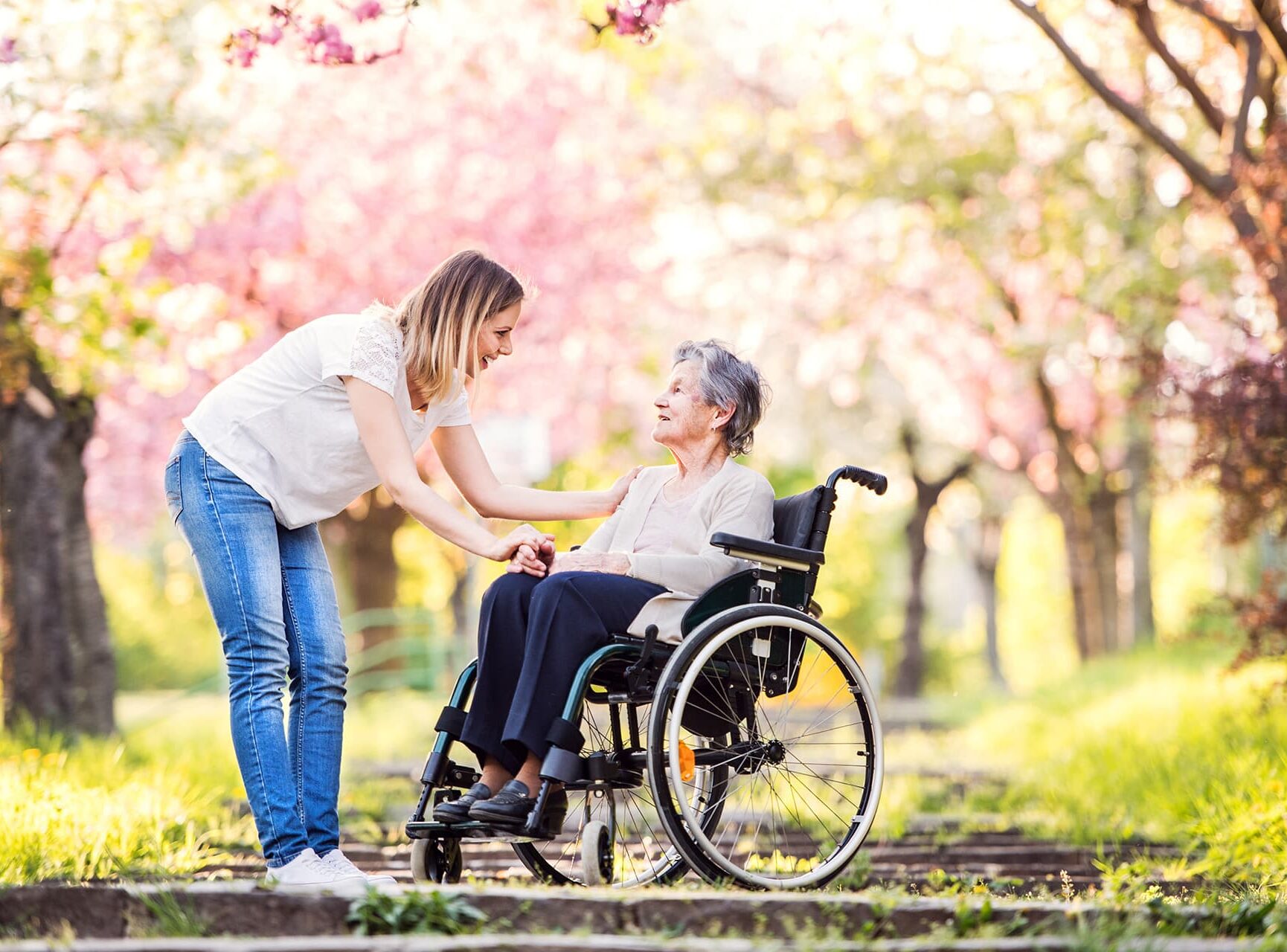 Eine alte Dame im Rollstuhl ist mit Pflegerin unterwegs im Park