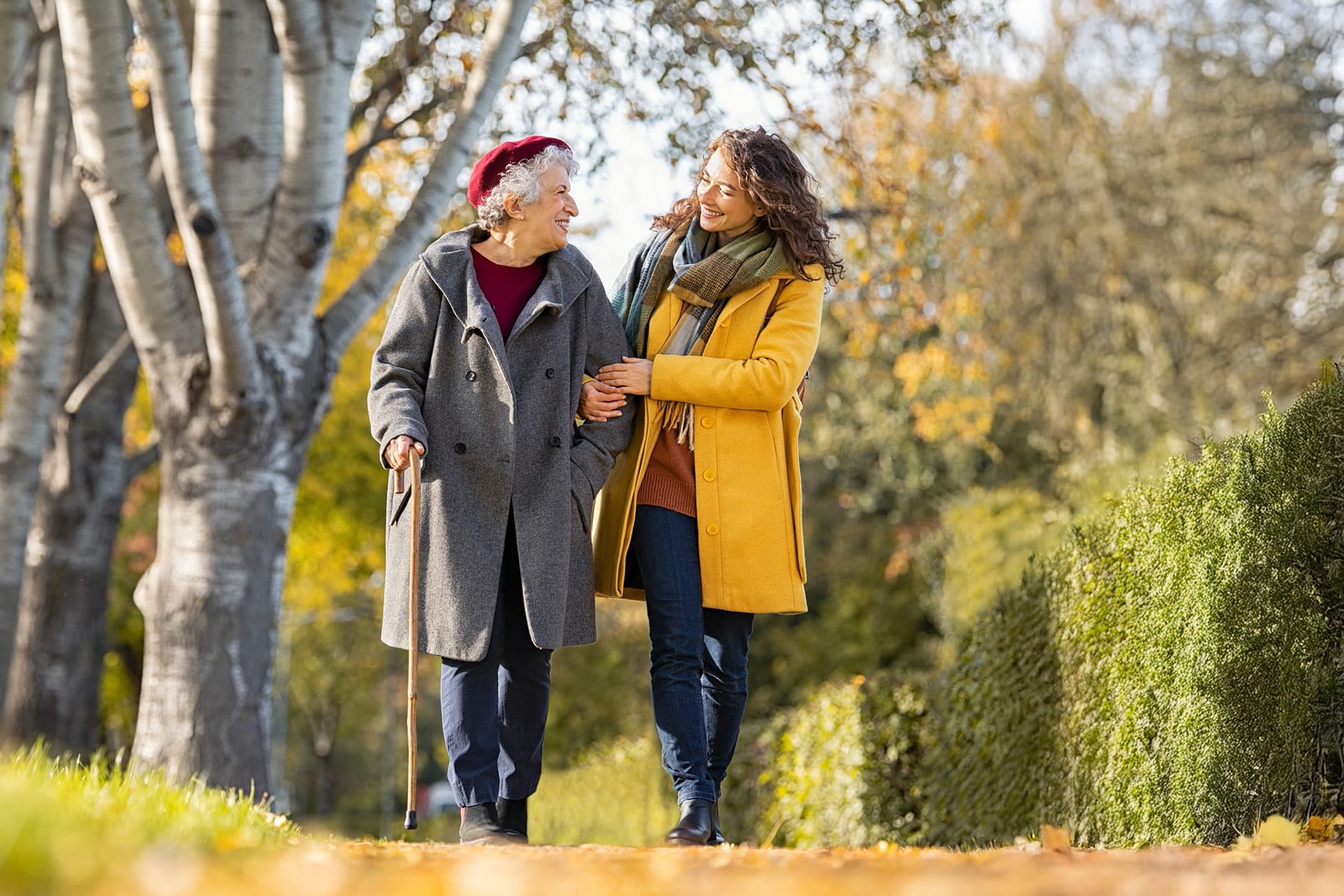 Ältere Frau mit Gehstock und eine freundliche Pflegekraft gehen im Park spazieren.