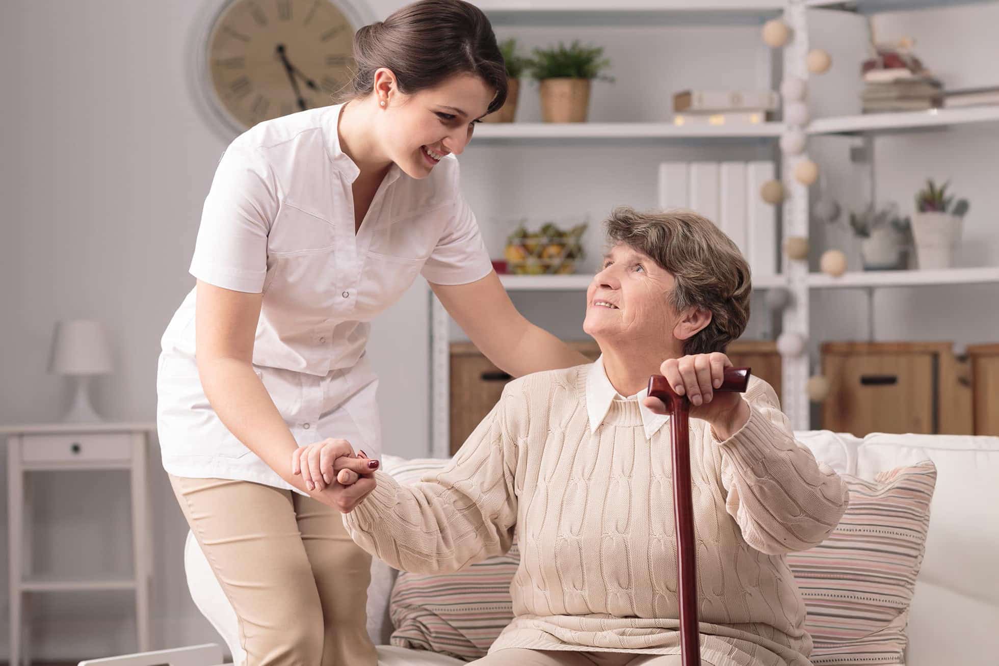 Pflegerin hilft alter Frau mit Kruecke beim Aufstehen